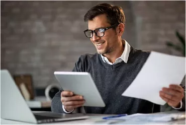 Man Smiling While Holding a Tablet and Papers While Looking at the Laptop